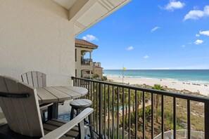 Un balcone con tavolo e sedie che si affaccia su una spiaggia, con una bandiera, palme e un edificio sullo sfondo.