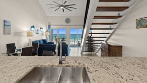 A modern kitchen with a marble countertop and stainless steel sink, a dining area with a table and chairs, a blue sofa, and a staircase leading to an upper level. - Beautiful 4th FL Condo, Gulf Front, Seasonal Beach Service (Miramar Beach)