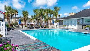 A swimming pool with lounge chairs and palm trees, surrounded by buildings. - 𝐏𝐚𝐢𝐱 𝐁𝐥𝐞𝐮 𝐓𝐨𝐨 Coastal Condo| Pool |Pets (Galveston)