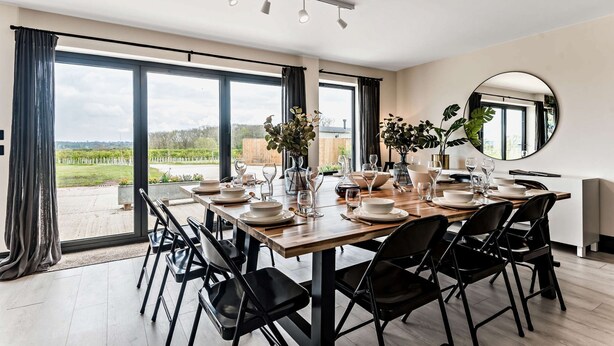 A dining area with a large table set for six, surrounded by black chairs, large windows with curtains, and a view of a garden. - Large 6 bedroom luxury holiday home in the Cotswolds with a hot tub - Folly View (Faringdon)