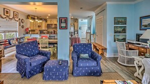 Ein Wohnzimmer mit zwei blauen Sesseln, einem Holztisch und einem weißen Sessel. Es gibt eine Essecke mit einem roten Tisch und Stühlen, und im Hintergrund eine Küche.