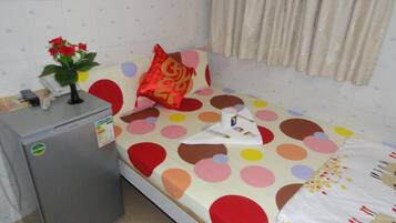 A small, single-bed room with a polka dot bedspread, a red pillow, a small fridge, and a plant on the nightstand.