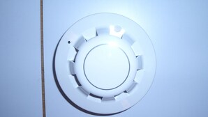 Un oggetto circolare, bianco e metallico, con un foro centrale e fessure circostanti, probabilmente parte di un meccanismo per porte o finestre.