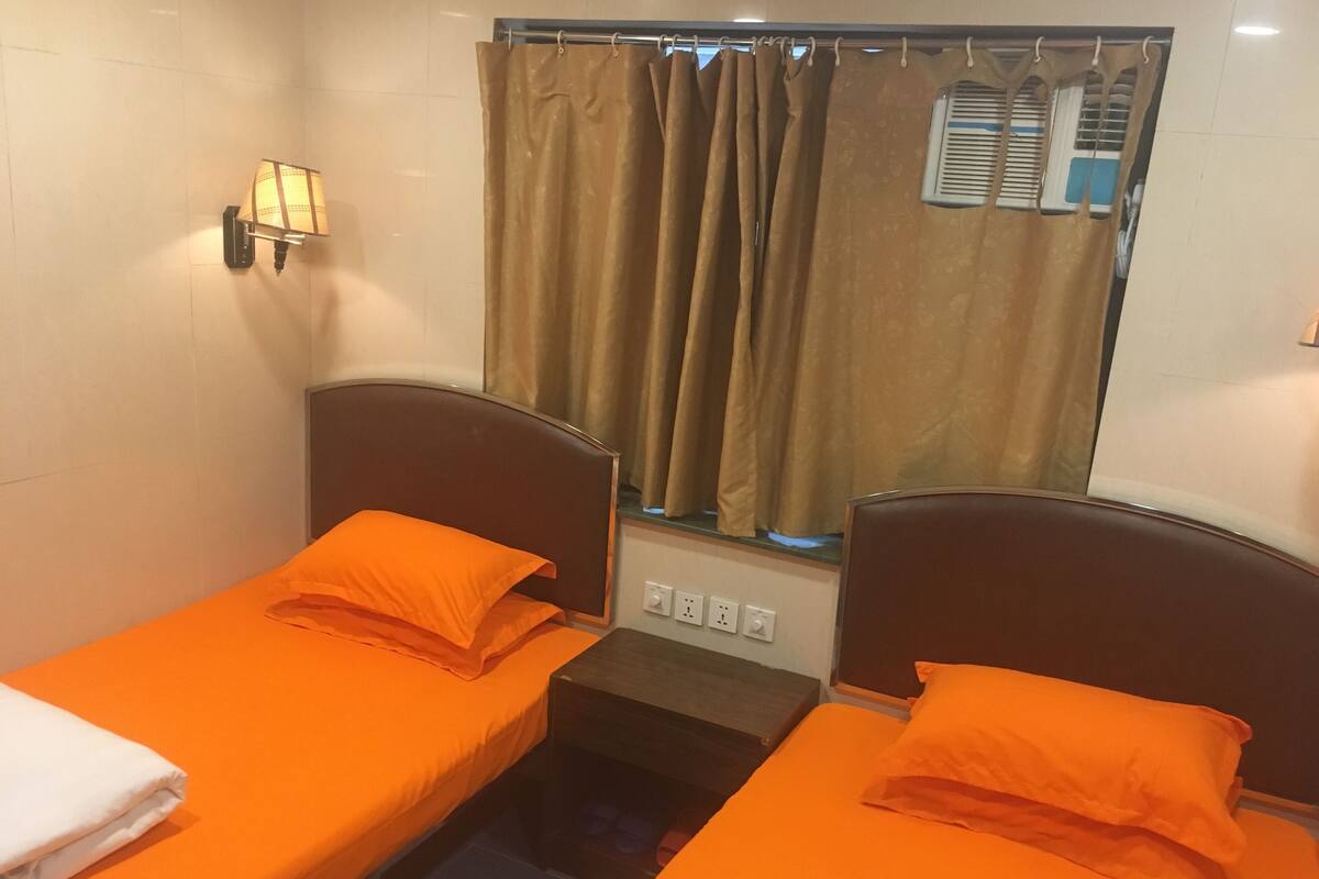 Une petite chambre avec deux lits simples, chacun recouvert d’une couverture orange assortie à des coussins. Un appliques mural est fixée sur le mur à gauche, et il y a une fenêtre avec des rideaux.