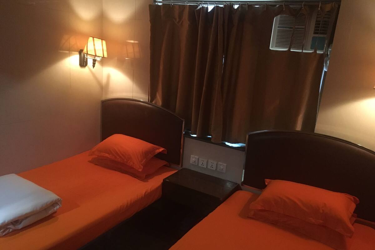 Une chambre avec deux lits simples, chacun étant équipé d’un oreiller et d’une couverture. Une table de chevet avec une lampe se trouve entre les deux lits.