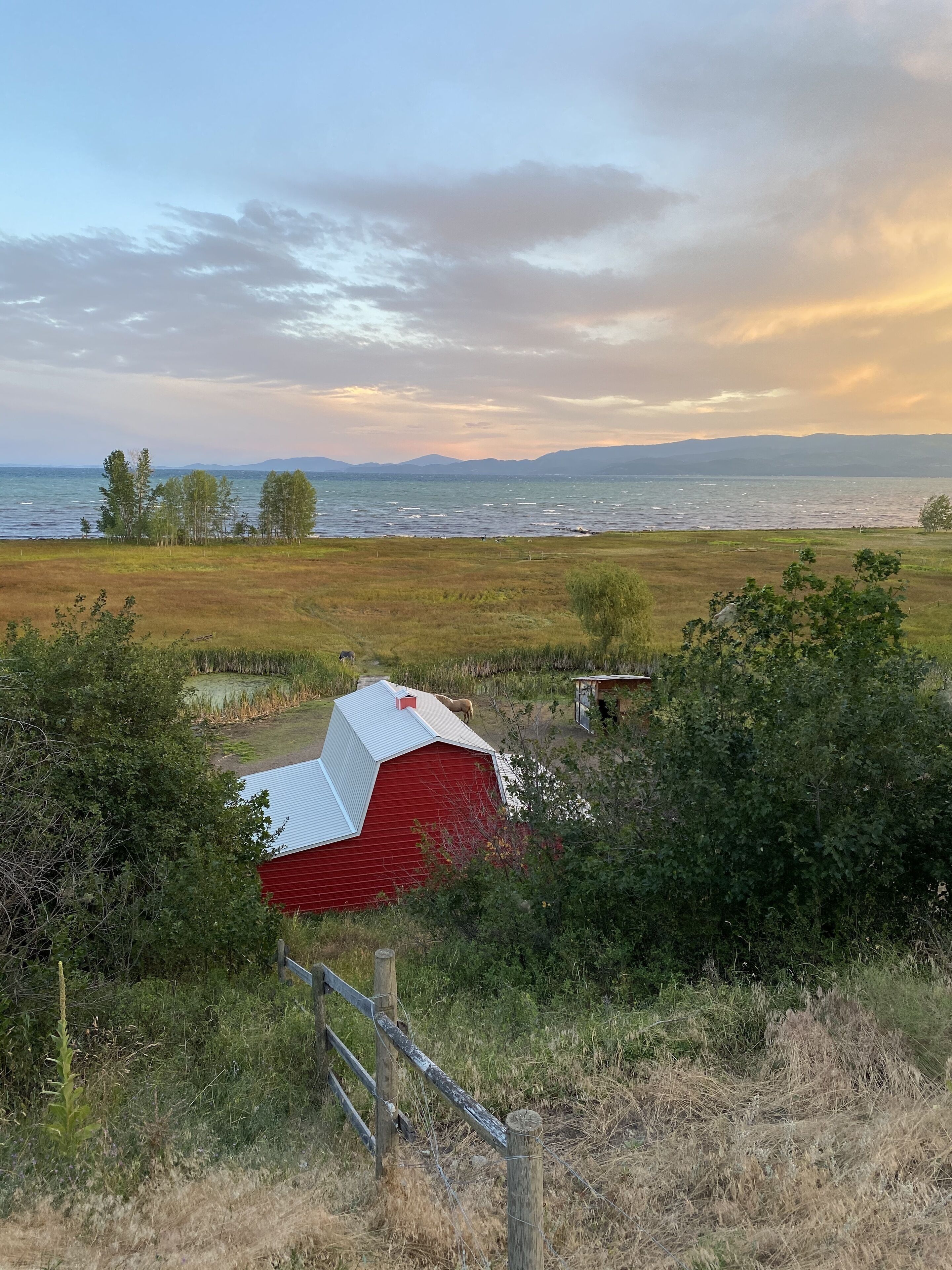 A red barn with a metal roof in a rural setting, surrounded by fields and trees, with a lake and mountains in the background.