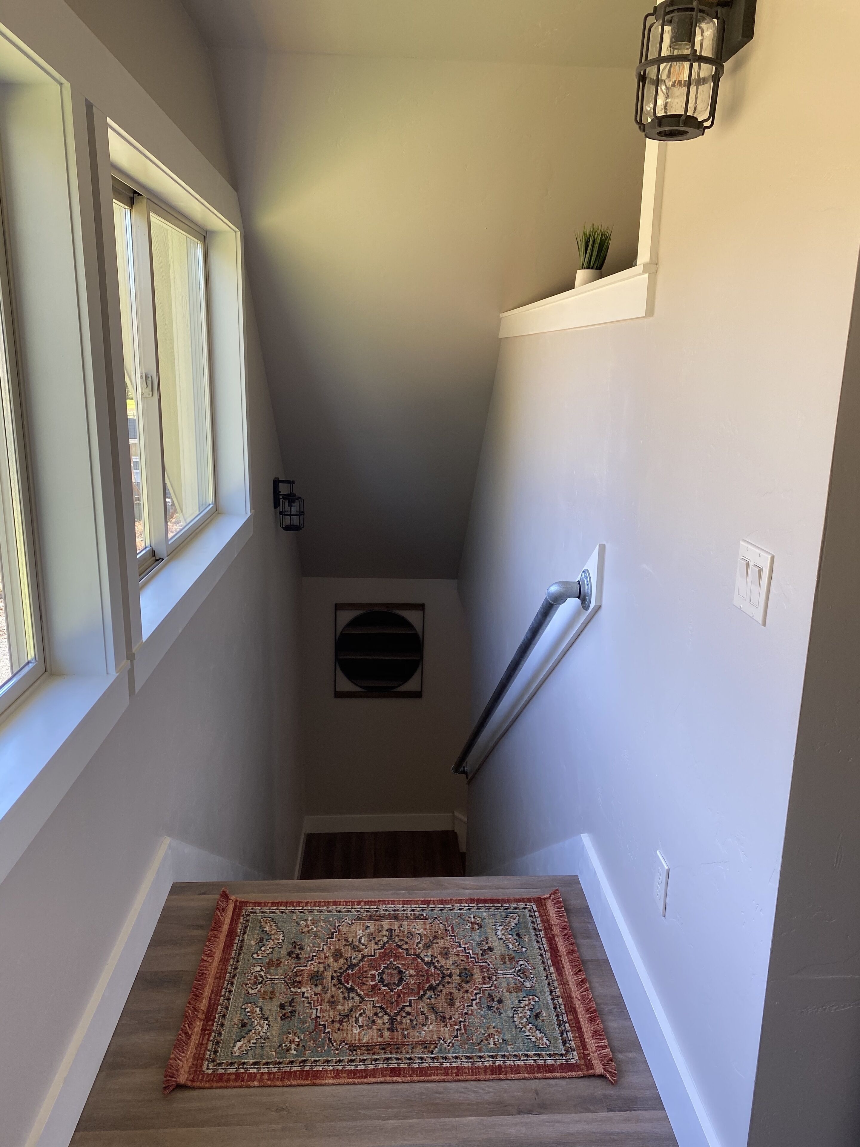 Un escalier étroit avec une rampe en bois, un tapis à motifs au bas de l’escalier et un luminaire fixé au mur.