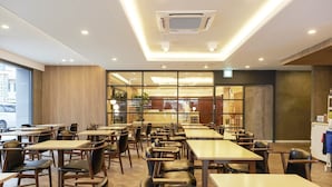 Un intérieur de restaurant moderne, avec de nombreuses tables et chaises, un appareil de climatisation fixé au plafond et de grandes fenêtres offrant une vue sur la rue.