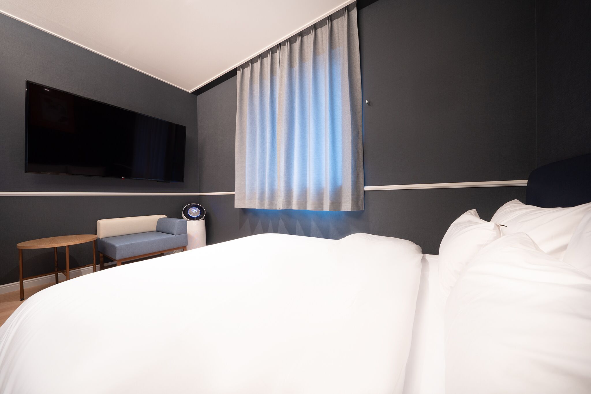 Et moderne hotelværelse med en stor seng, et fladskærms-tv monteret på væggen, et lille rundt bord, en blå sofa og et vindue med gardiner.