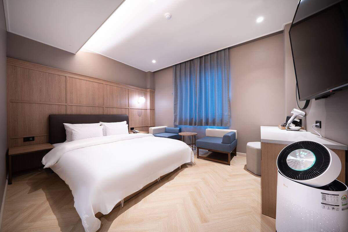 Une chambre d’hôtel moderne dotée d’un grand lit, d’un coin salon avec un canapé et des fauteuils, d’une petite table, d’un téléviseur à écran plat et d’un purificateur d’air sur pied.