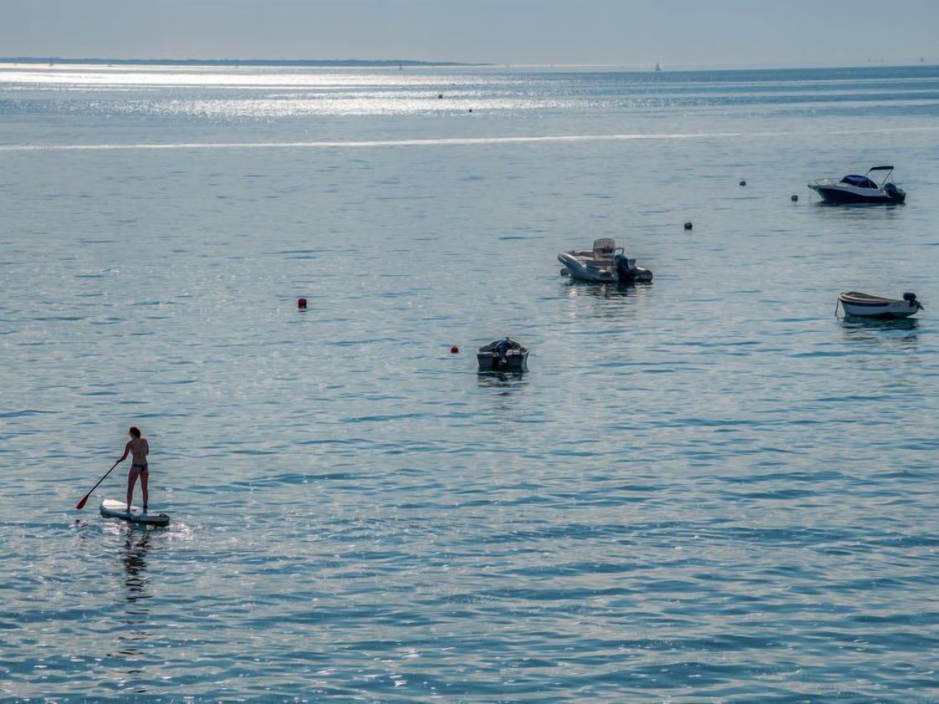 Una persona fa paddleboarding su acqua calma, con diverse barche e boe sullo sfondo.