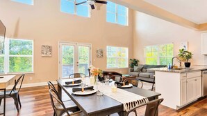 A spacious dining area with a long table, chairs, and a ceiling fan. There are large windows with blinds, a kitchen area with a sink and cabinets, and a gray sofa in the background. - Hot Tub! City Side Comfort in Missionary Ridge (Chattanooga)