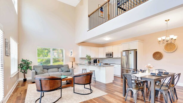A spacious living area with a grey sofa, wooden coffee table, and a dining table with metal chairs. A kitchen with white cabinets and stainless steel appliances is visible in the background. - Hot Tub! City Side Comfort in Missionary Ridge (Chattanooga)
