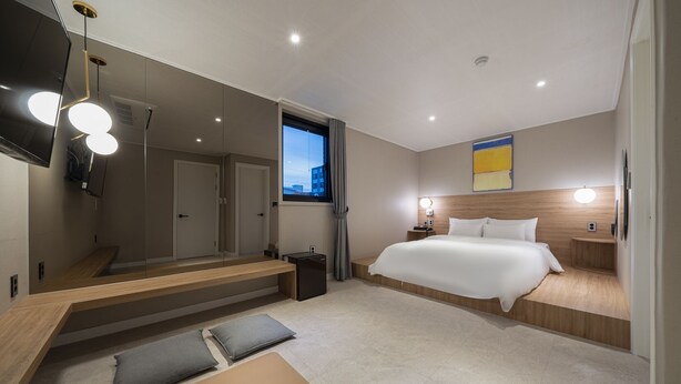 A modern hotel room with a large bed, a wooden bench, a small table, a mirror, a TV, and a window with curtains. - Goyo37 Hotel Osan by Anook (Osan)