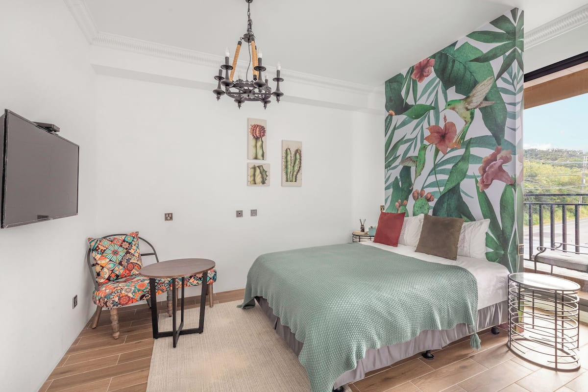 Quarto com cama, poltrona, mesa pequena, lustre e parede com mural floral.