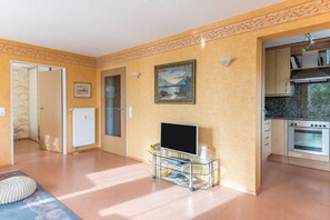 A living room with a television on a glass stand, a painting on the wall, and a kitchen area with a stove.