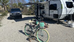 A recreational vehicle with a canopy, a bicycle, and a dog in a gravel area with palm trees and a truck in the background. - Incommunicado Gulf Front Private RV Lot (Port St. Joe)
