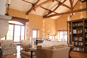 Un ampio soggiorno con soffitto alto, pareti in pietra e travi in legno a vista. Sono presenti diverse zone relax, una libreria e un caminetto.