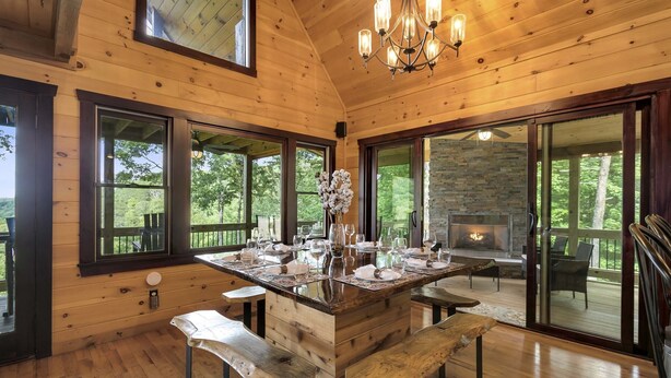 Een houten chalet binnen met een grote eettafel, banken, een open haard en grote ramen met uitzicht op de natuur.