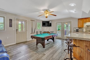 A pool table in a room with a ceiling fan, a television mounted on the wall, and a kitchen area with wooden cabinets. - Break Away - sleeps 12, kayaks, SUP's fire pit, games galore! Great for kids and adults! (Moneta)
