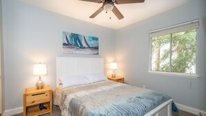 A bedroom with a bed, two bedside tables with lamps, a ceiling fan, a window with a view of trees, and a painting on the wall.