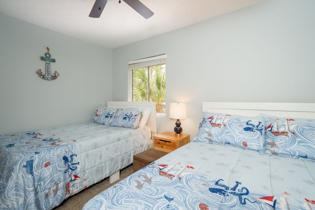A bedroom with two beds, a ceiling fan, a window with a view of palm trees, and a decorative anchor on the wall.
