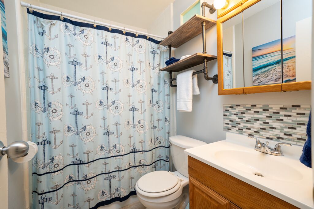 A bathroom with a nautical-themed shower curtain, a white toilet, a wooden vanity with a sink, and a mirror above the vanity.
