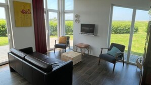 A modern living room with a black leather sofa, a wooden coffee table, and a television on the wall. Large windows offer a view of a green landscape.