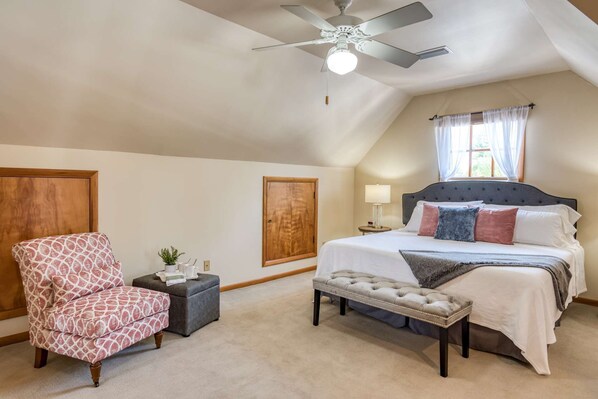 A bedroom with a large bed, a chair, a nightstand, a ceiling fan, and a window with curtains.