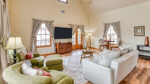 A spacious living room with a high ceiling, a flat-screen TV mounted on the wall, a wooden coffee table, a sofa, and two armchairs.