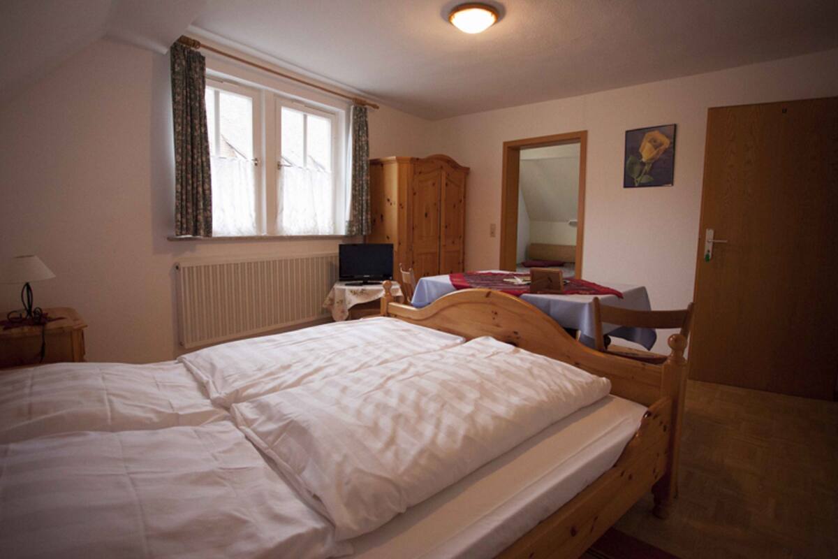 A bedroom with a double bed, a wooden wardrobe, a desk with a computer, and a window with curtains.