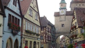 Una plaza histórica en un pueblo europeo, con edificios tradicionales de entramado de madera, una torre del reloj destacada y calles empedradas.