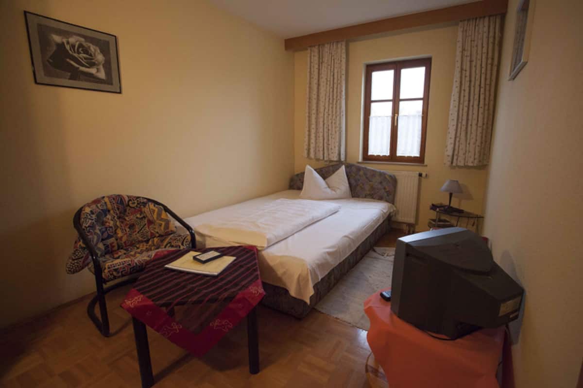 A small, neatly arranged bedroom with a single bed, a chair, a table, and a television.
