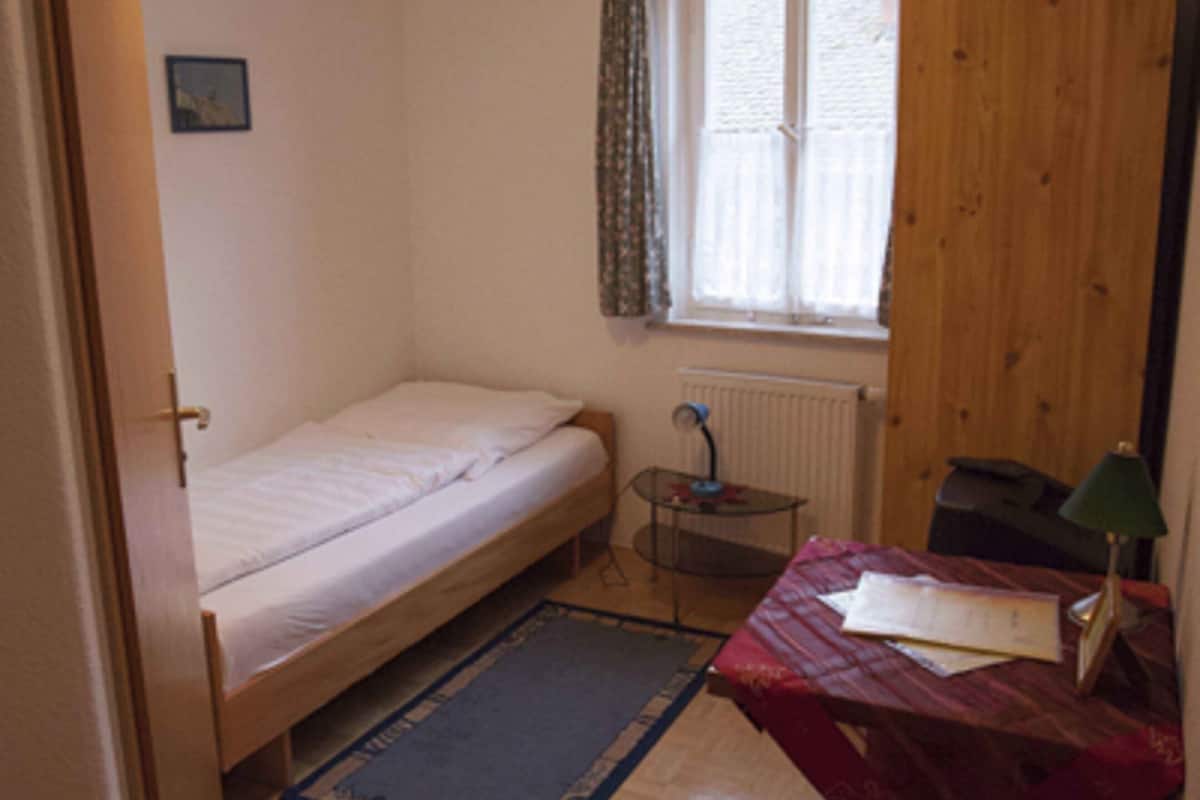 A small, single-bed room with a wooden floor, a desk with a chair, a radiator, and a window with curtains.