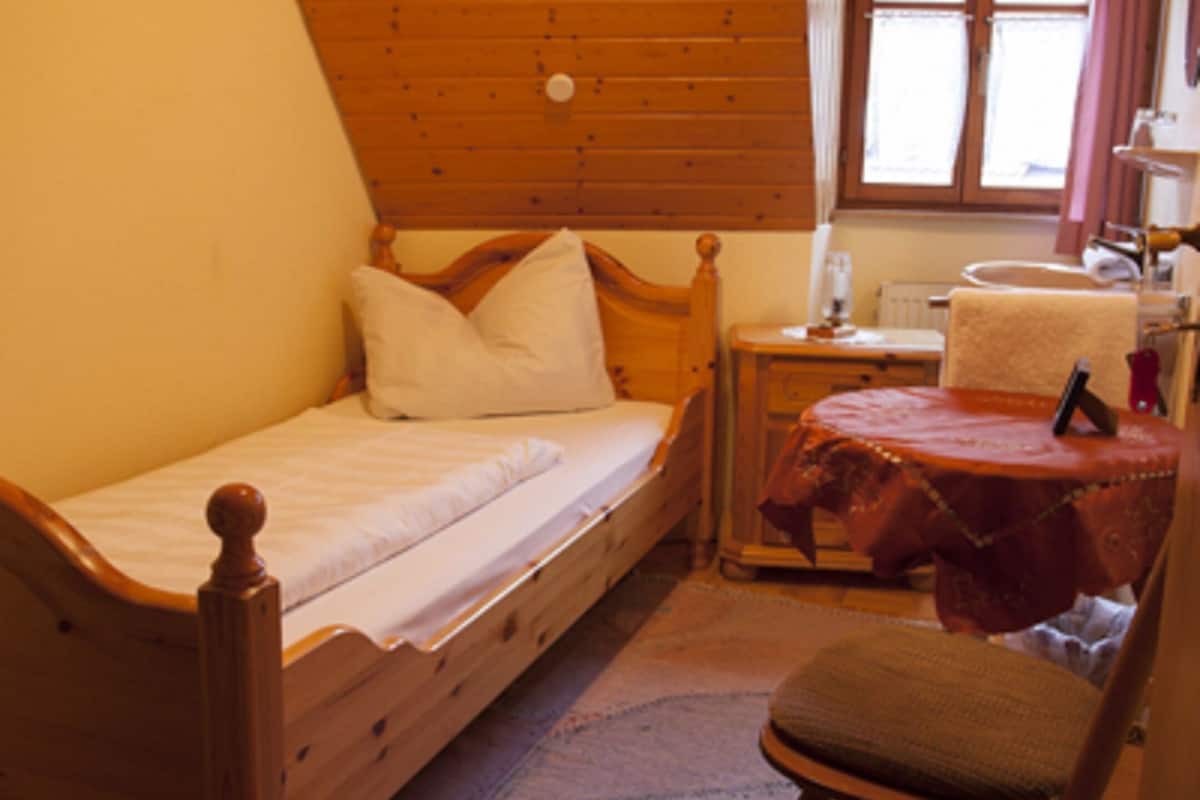 A small, cozy room with a wooden bed, a chair, a small table with a tablecloth, and a window with curtains.