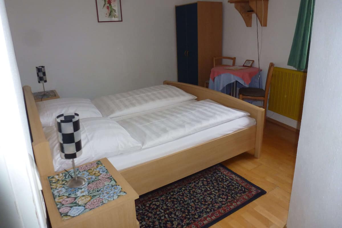 A single bed with a wooden frame, a bedside table with a floral-patterned tablecloth, a small chair, a radiator, and a television mounted on the wall.