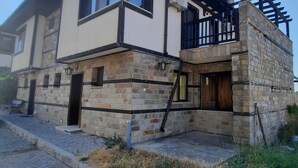 A two-story house with a stone facade, a balcony with a black railing, and a clear sky. - Beautiful 2-bed Villa in Cholakova Cheshma (Kosharitsa)