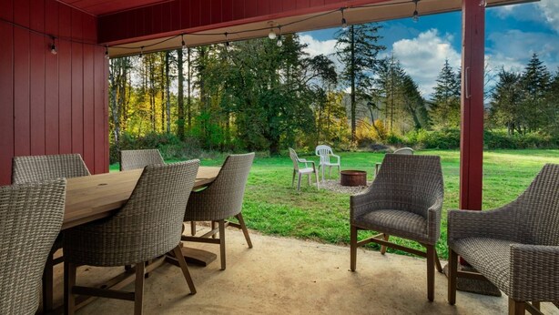 Een terras buiten met rieten meubilair en een houten tafel, omgeven door een rode constructie met uitzicht op een bebost gebied.
