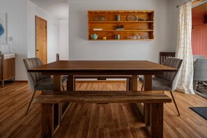 A dining area with a wooden table and chairs, a shelf with kitchen items, and a view of a red wall and a patterned curtain.