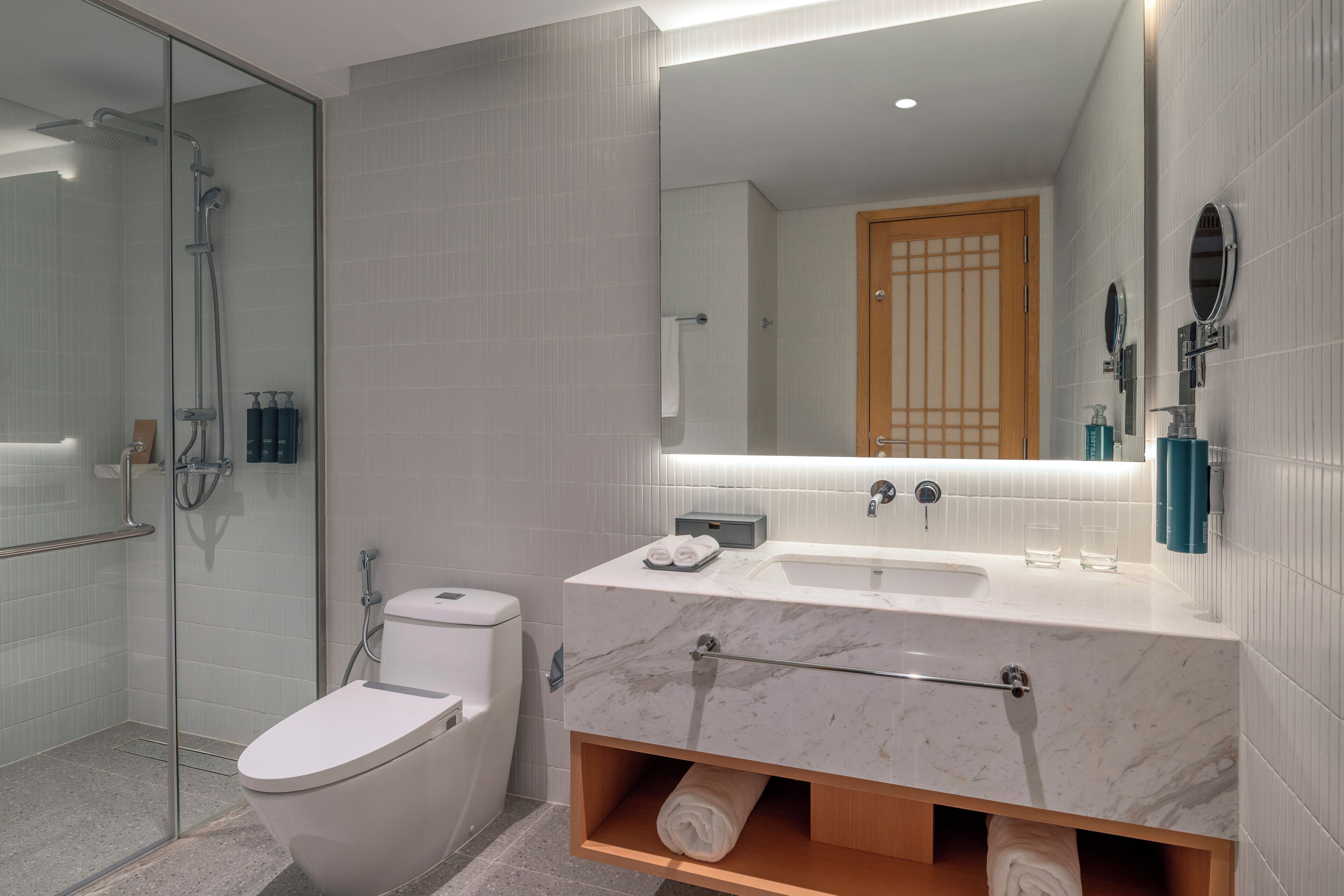 Un baño moderno con ducha de vidrio, inodoro blanco, lavamanos de mármol y tocador de madera.