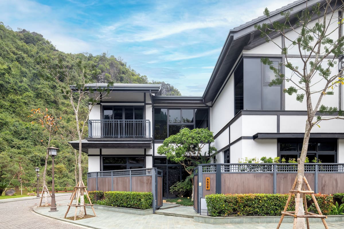 Um edifício moderno com varanda, entrada com portão e uma encosta verdejante ao fundo.