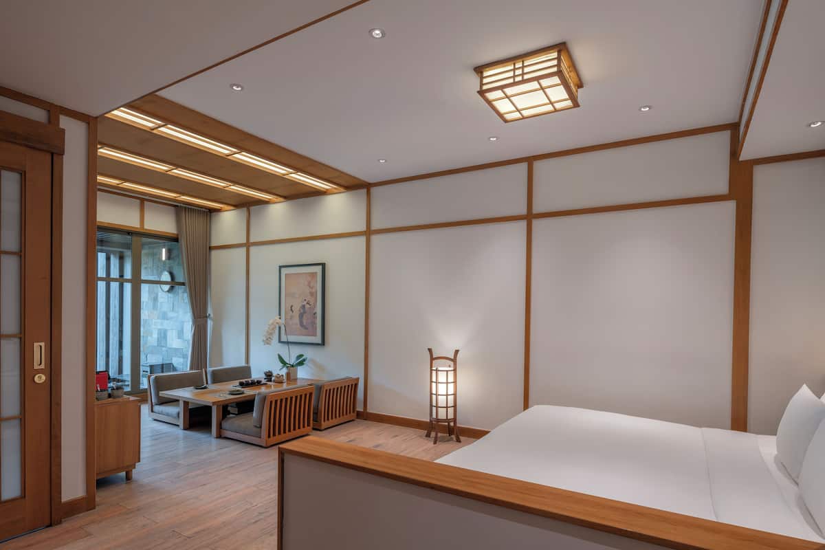 Quarto de hotel moderno com piso de madeira, uma cama, uma escrivaninha, uma cadeira, uma mesinha de apoio, uma luminária e uma janela com cortinas.