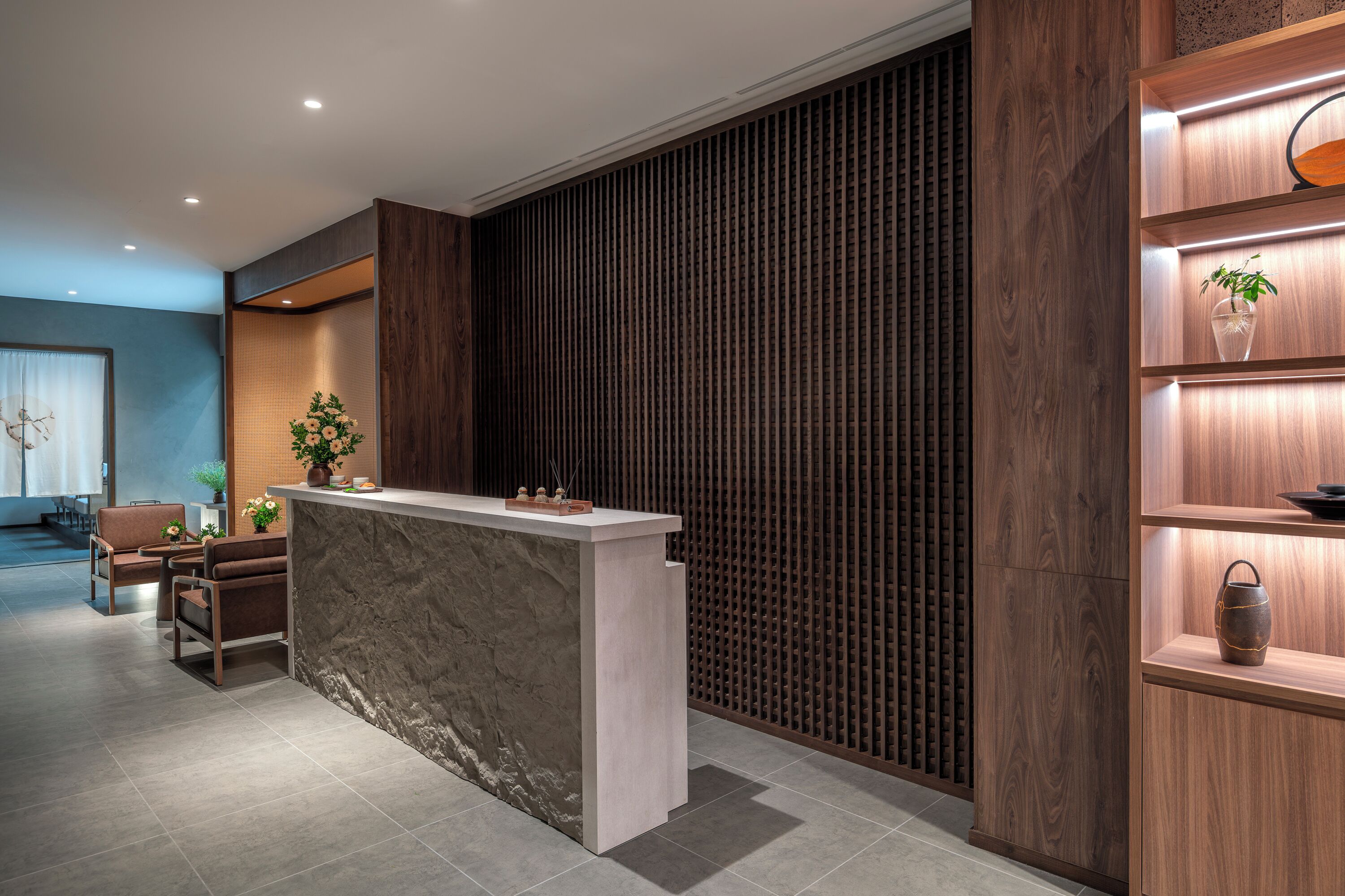 Un mostrador de recepción con encimera de mármol, estanterías de madera con objetos decorativos y un área de descanso con sillas y una mesa.
