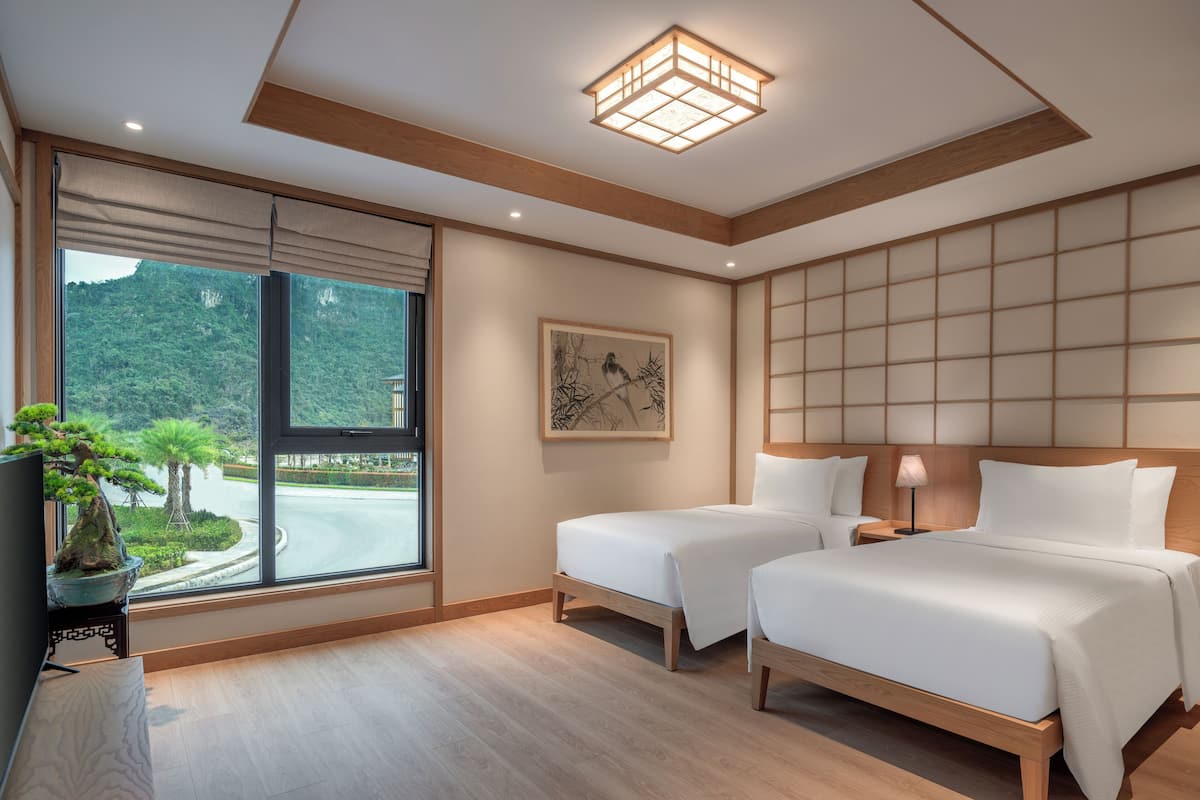 Quarto de hotel com duas camas, uma janela grande com vista para vegetação, um candeeiro no teto e uma obra de arte emoldurada na parede.