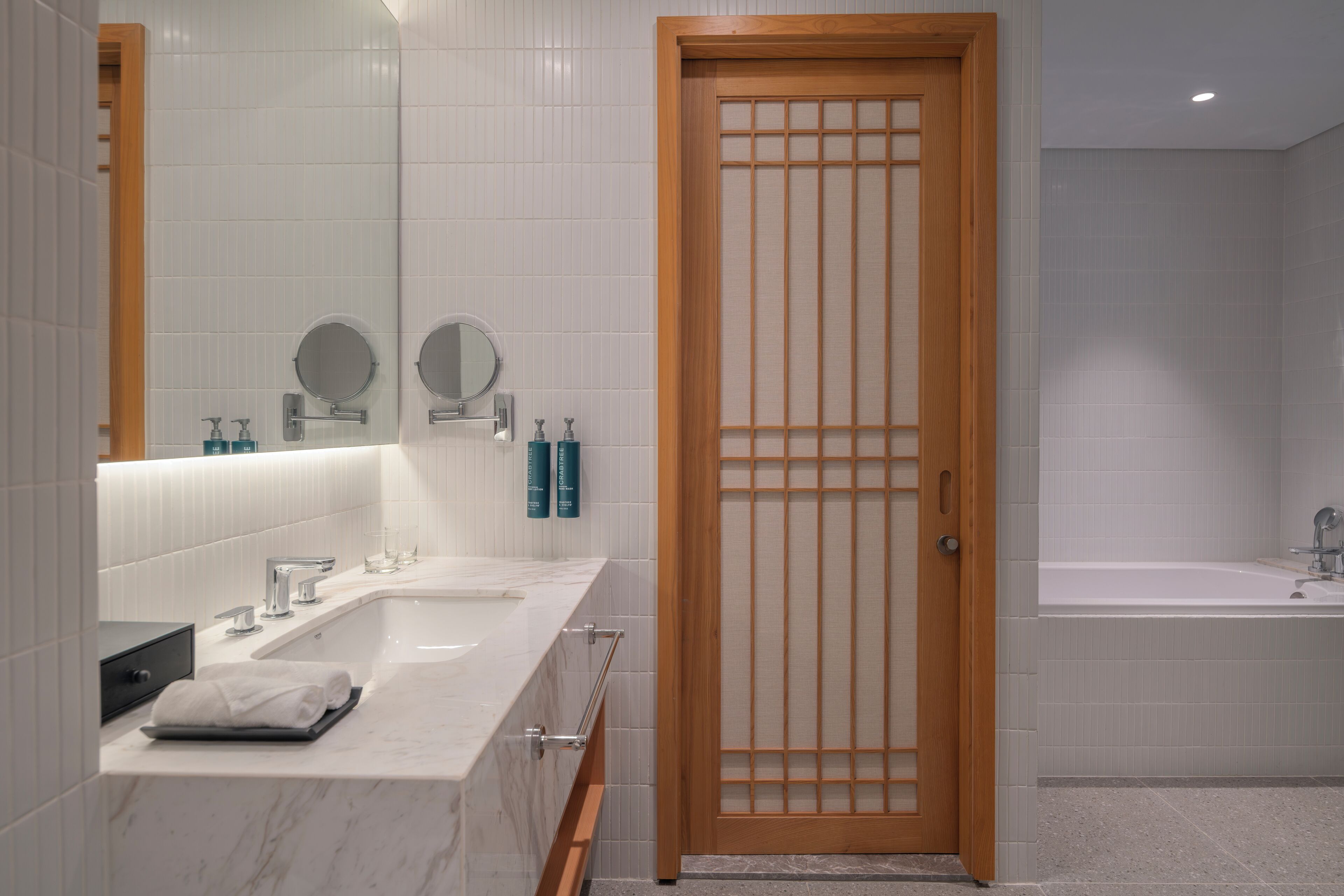 A modern bathroom with a double sink, a large mirror, a wooden door, and a bathtub.