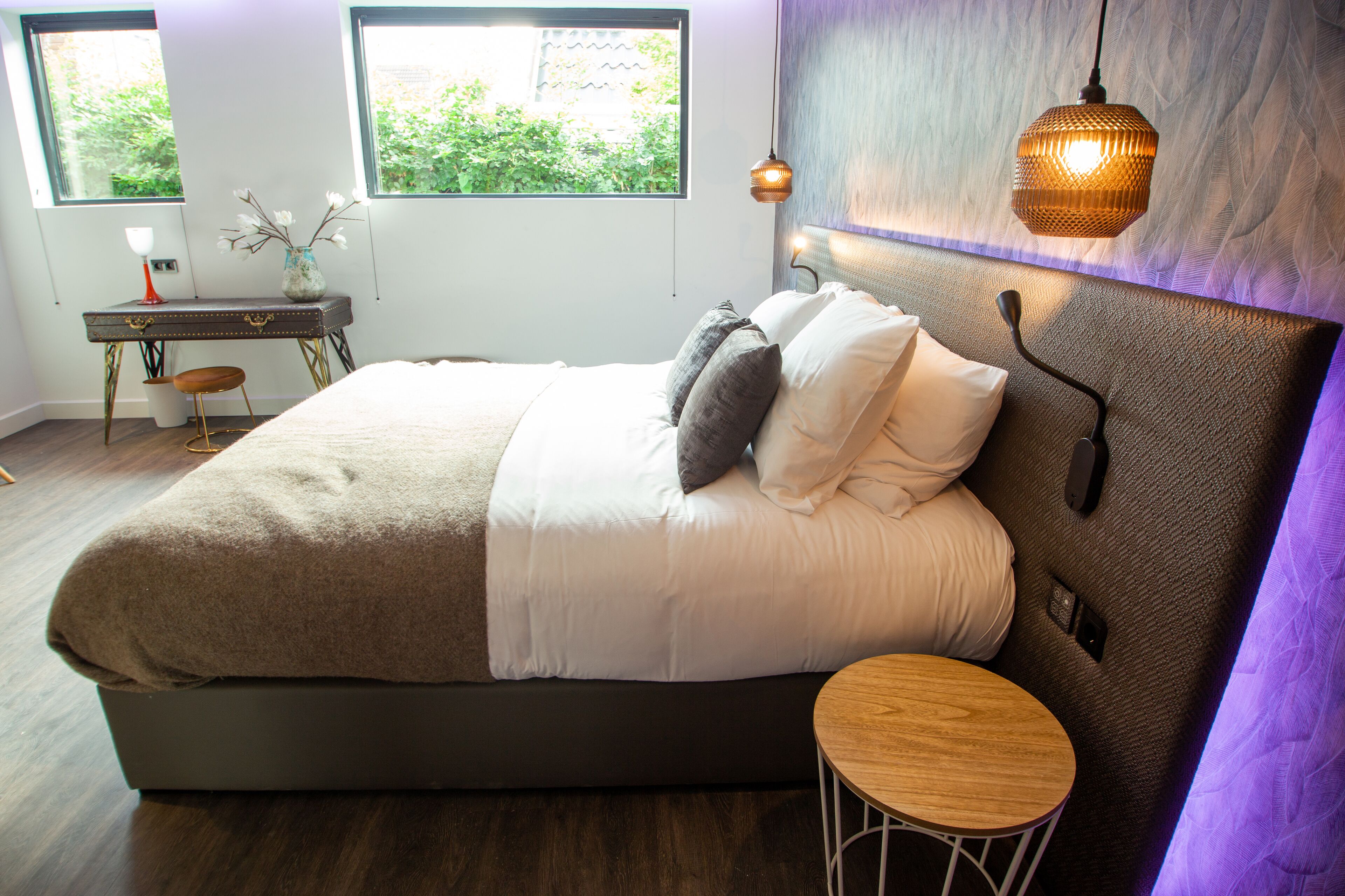 Een slaapkamer met een groot bed, nachtkastje en een klein krukje. Er zijn ramen met uitzicht op groen.