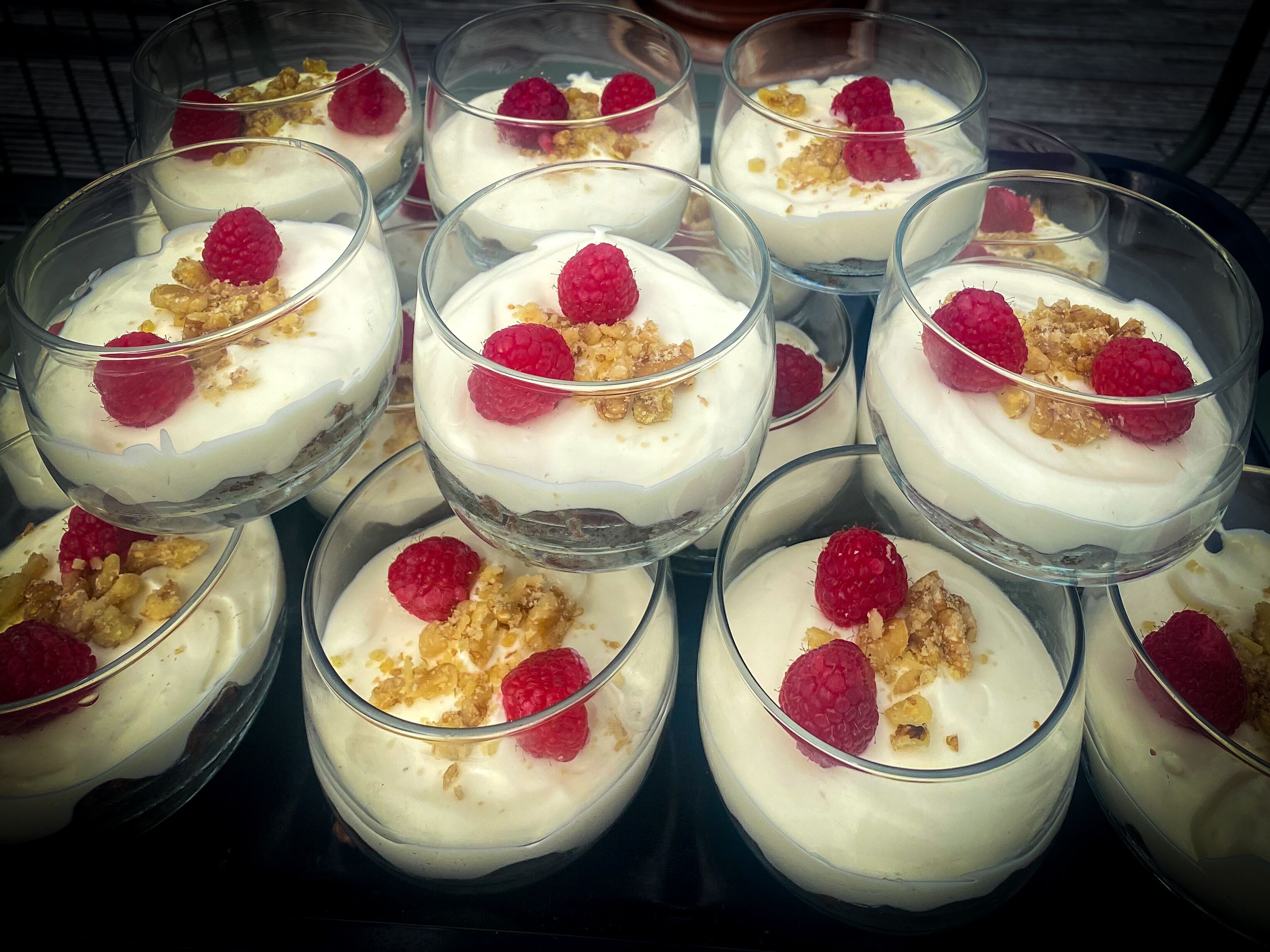 Een schaal met dessertbekers, met witte romige laagjes, afgetopt met frambozen en gehakte noten.
