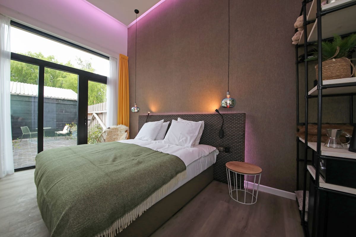 A modern bedroom with a large bed, a small stool, a shelf with decorative items, and a view of the outdoors through a sliding door.