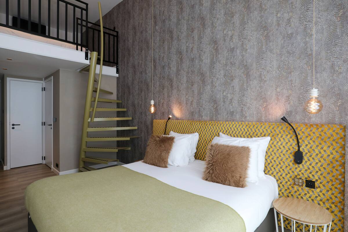 A bedroom with a bed, a small table, a staircase, and a wall with a patterned design.