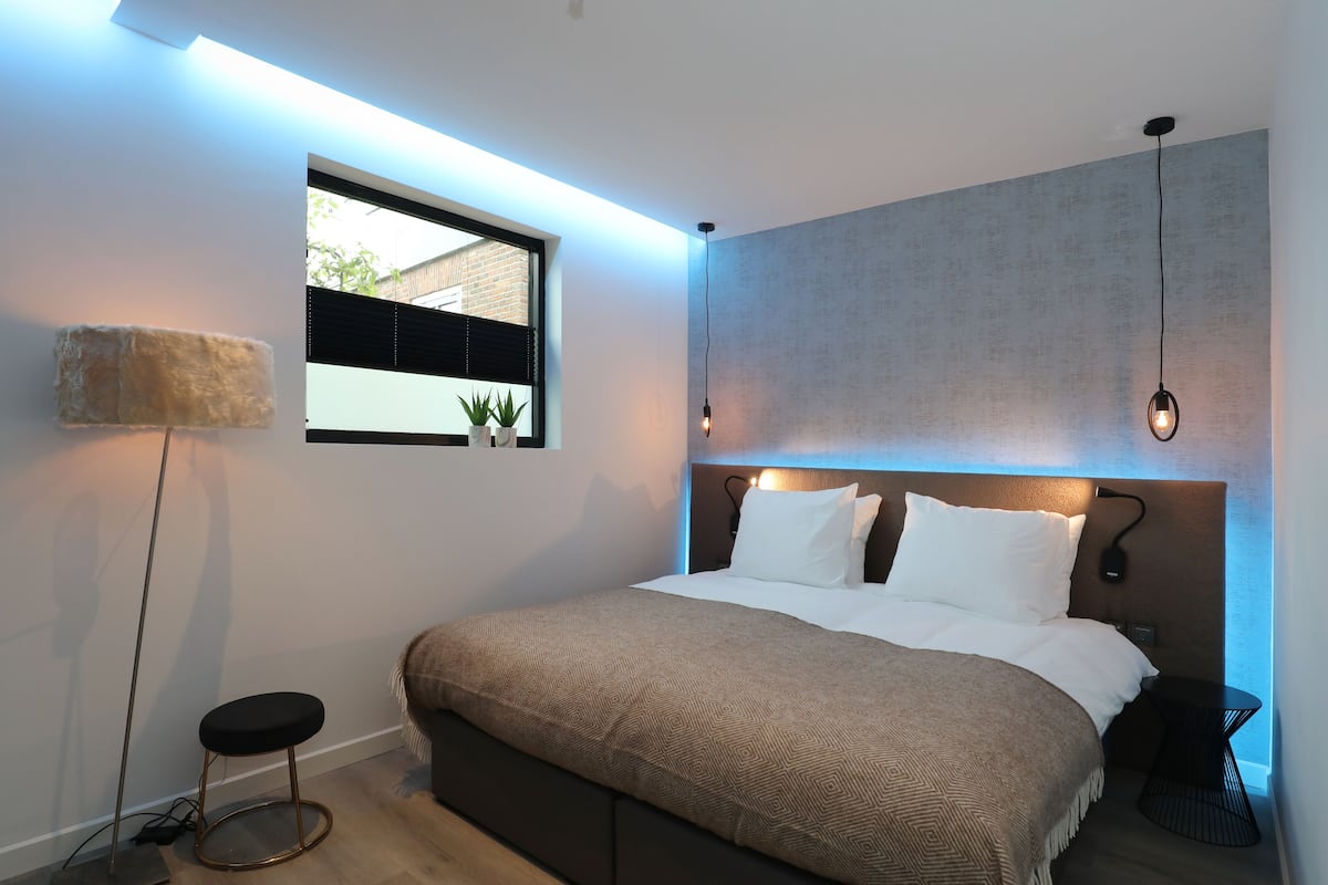 A modern bedroom with a large bed, a small stool, a side table, and a window with blinds.
